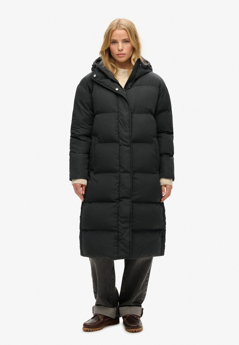 Langer schwarzer Daunenmantel mit Kapuze, ausgestattet mit einem Reißverschluss und Druckknopfverschluss, großen gesteppten Abschnitten und seitlichen Taschen. Kombiniert mit grauen Jeans und Stiefeln.