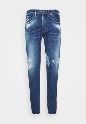 Jean en denim bleu avec une coupe slim, des zones délavées et des patchs usés sur les cuisses et les genoux.