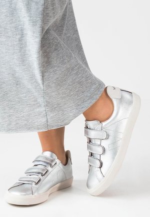 Sølvmetallic sneakers med tre Velcro-stropper, rund tå og tekstureret overflade, parret med et gråt afslappet-fit tøj.