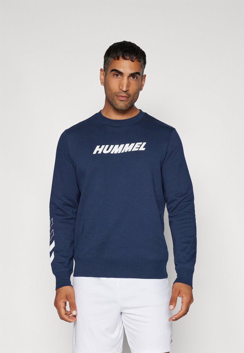 Mørkeblå sweatshirt lavet af bomuld med lange ærmer, har et hvidt "HUMMEL" logo på fronten og et geometrisk mønster langs ærmet.
