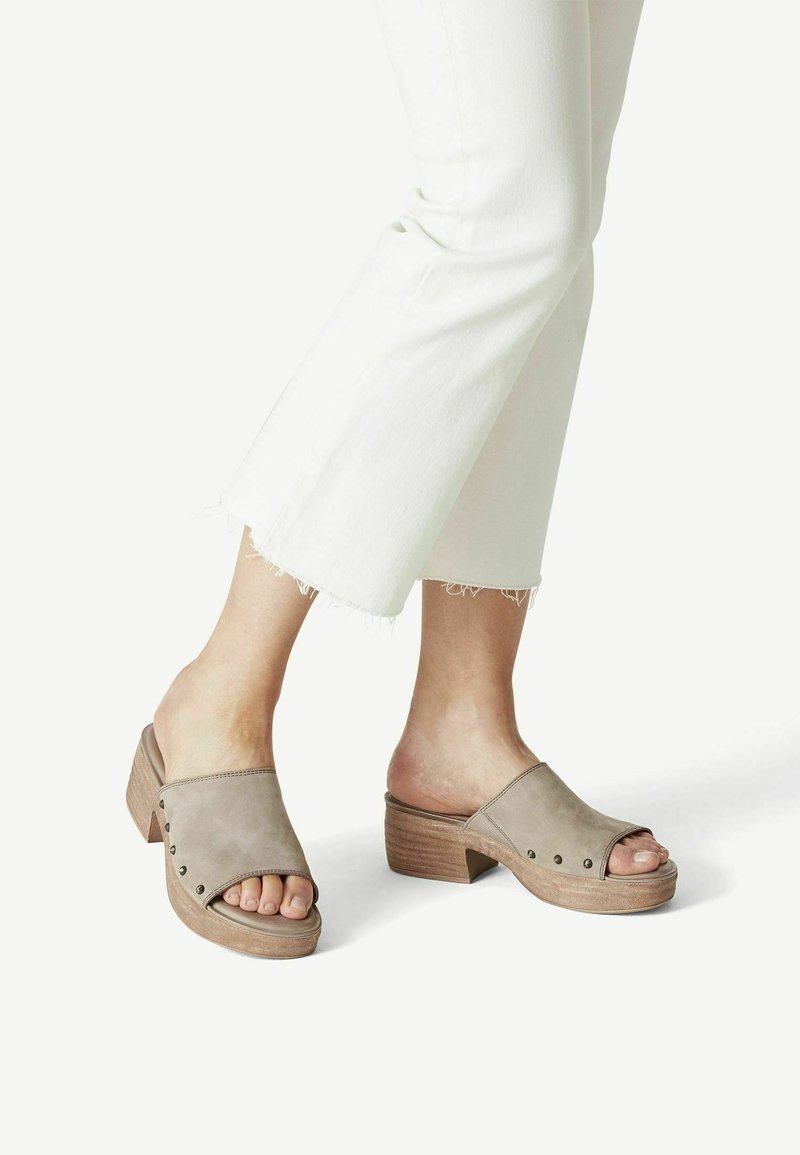 Mules beiges à bout ouvert avec une finition en daim lisse, ornés d'accents à rivets sur les côtés et d'un talon en bois empilé. Portés avec un pantalon blanc court.