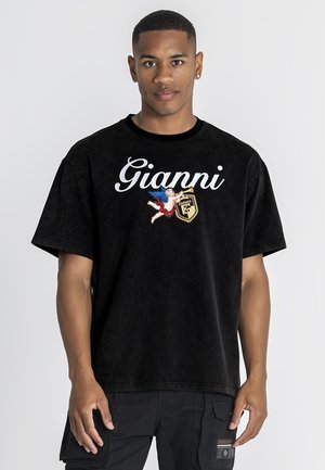 Jeune homme portant un T-shirt noir oversize avec le texte "Gianni", un chérubin et un dessin de bouclier, associé à un pantalon cargo noir.