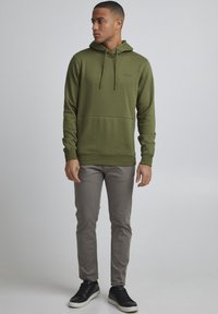 Haut à capuche vert olive avec poche avant, cordon de serrage réglable et marque discrète ; associé à un pantalon gris clair et des baskets noires.