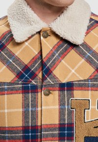 Plaid shirt jacket in tan, red, and blue, with a fluffy cream collar, detailed stitching, and a patch featuring a letter in textured fabric.