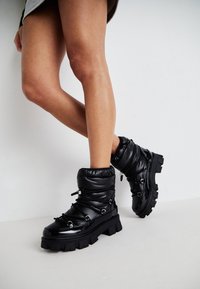 Black padded ankle boots with a chunky sole, featuring a textured upper, elastic drawstring, and metallic accents.