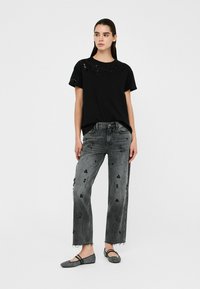 Schwarzes T-Shirt mit floralen Verzierungen, kombiniert mit ausgewaschenen grauen Jeans mit dekorativen Akzenten. Model trägt flache Schuhe.