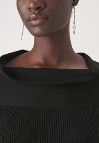 Fekete kötött ruha széles, hajtott dekoltárral. A derék részen texturált minta található, függőleges vonalakkal és geometriai díszítéssel. Ezüst csepp fülbevalók.