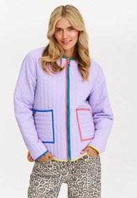 Kobieta nosząca lawendową pikowaną kurtkę z kolorowymi kieszeniami i zamkiem, w zestawieniu z spodniami w panterkę, stojąca na białym tle.