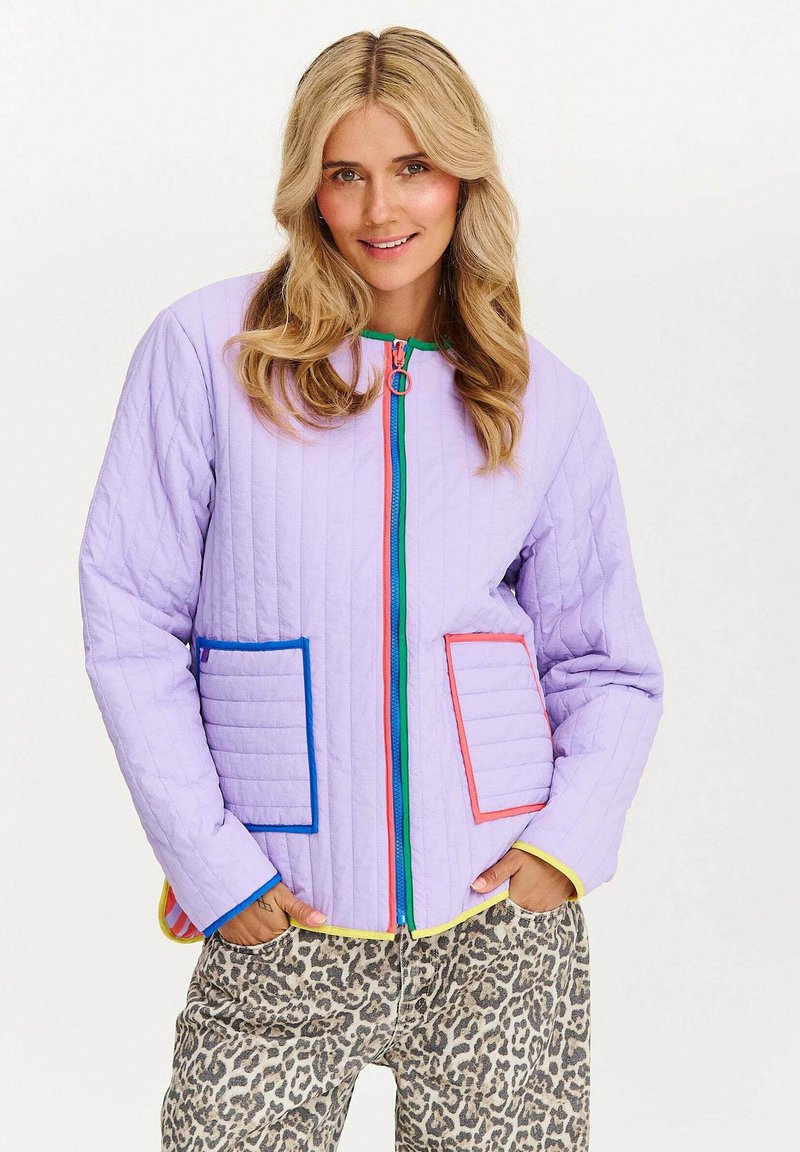 Kobieta nosząca lawendową pikowaną kurtkę z kolorowymi kieszeniami i zamkiem, w zestawieniu z spodniami w panterkę, stojąca na białym tle.