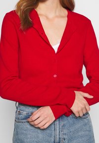 Rote Strickjacke mit Knopfleiste, gerippter Textur und einem Kragen. Getragen über einem weißen Oberteil und kombiniert mit hochtaillierten blauen Jeans.