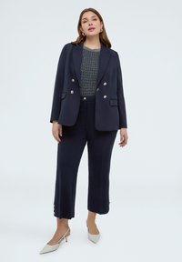 Frau trägt marineblauen Blazer mit silbernen Knöpfen, passende cropped Hose, graues strukturiertes Oberteil, weiße Slingback-Pumps, steht vor einfarbigem Hintergrund.