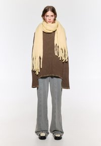 Braun langärmliges Strickoberteil, kombiniert mit grauen weit geschnittenen Hosen. Ein großer fransenbesetzter gelber Schal sorgt für Textur und kontrastiert mit den Farben des Outfits.