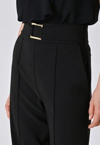 Person trägt schwarze hochgeschnittene Hose mit einem breiten Bund und einer goldenen rechteckigen Schnalle, kombiniert mit einem schwarzen Oberteil.