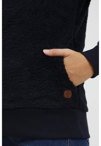 Pullover in pile navy con superficie strutturata e morbida. Presenta una tasca frontale e polsini a coste. Applicazione del logo marrone nell'orlo.