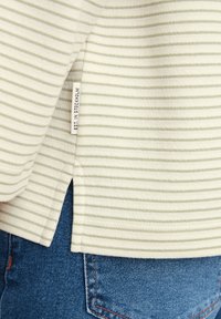 Haut à manches longues rayé en crème et vert clair, présentant une fente sur le côté et une étiquette intitulée "EST. À STOCKHOLM." Associé à un jean en denim bleu.