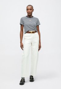 Žena stojaca v čierno-bielom pruhovanom tričku, bielych nohaviciach s vysokým pásom, hnedým pásom, sivých ponožkách a čiernych polobotkách.