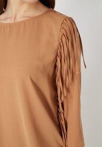 Top color beige con un escote redondo y flecos decorativos en las mangas, confeccionado con una tela suave y de textura agradable.