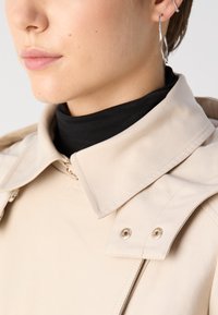 Beige trenchcoat met een grote kraag en drukknoopdetails, met een gladde textuur en subtiele stiksels. Zwarte coltrui zichtbaar eronder.