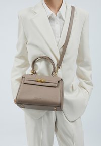 Graue Lederhandtasche mit strukturierter Form, goldfarbenen Beschlägen und abnehmbarem Schulterriemen, gehalten von einer Person in einem weißen Anzug.