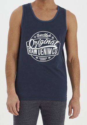 Homme portant un débardeur bleu marine avec le logo blanc « Original Raw Denim Co. », assorti à un pantalon gris foncé texturé.