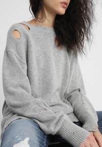 Pull tricot gris surdimensionné avec des détails déchirés prononcés sur les épaules et les poignets. Texture douce et accents côtelés à l'ourlet et aux manches.