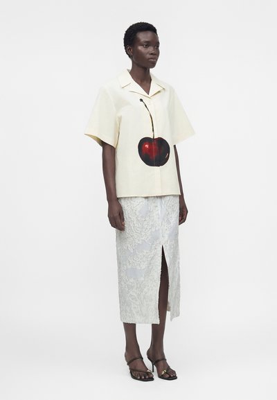 Γυναίκα που φοράει κρεμ κοντομάνικο πουκάμισο με μεγάλο σχέδιο κερασιού και λευκή φλοράλ δαντελένια φούστα με μπροστινή σχισμή και μαύρα σανδάλια με τακούνι.