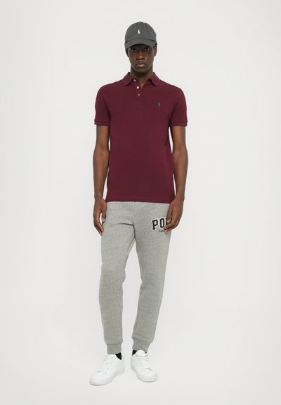 Bordo polo majica, sive trenirka hlače s 'Polo' natpisom, bijele tenisice i sivi kap. Uski kroj, ležeran atletski stil, minimalni detalji.