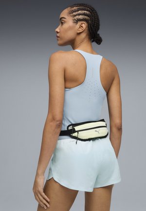 Mujer con el cabello trenzado, llevando una camiseta sin mangas azul claro y pantalones cortos, de espaldas, con una riñonera Puma crema y negra alrededor de la cintura.