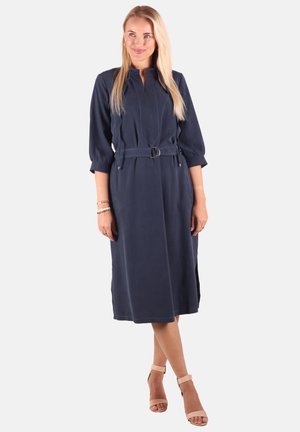 Blonde Frau trägt ein marineblaues Midikleid mit Gürtel, dreiviertel Ärmeln und beigen Knöchelriemenabsätzen, steht auf hellem Hintergrund.