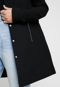 Chaqueta negra texturizada con botones a presión y un bolsillo con cremallera lateral. Combinada con vaqueros de mezclilla azul claro, mostrando un diseño de corte holgado.