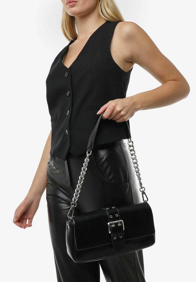 Borsa a tracolla in pelle nera con forma rettangolare, caratterizzata da un dettaglio a fibbia, una tracolla in catena e una superficie liscia. Indossata da una modella in abbigliamento nero.