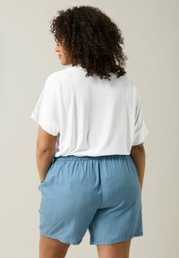 Camicia bianca a maniche corte abbinata a pantaloni corti azzurri chiari con vita elastica e tasche laterali, che mostrano una vestibilità rilassata e un tessuto morbido.