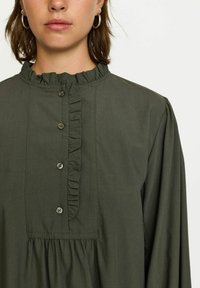 Blouse vert olive en tissu doux, avec un col volanté, une fermeture à bouton sur le devant, et une taille froncée avec des manches longues.