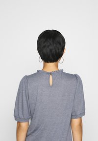 Top gris de manga corta con escote redondo, que presenta un borde con volantes y un detalle de ojal en la parte posterior. Tejido suave con ligera textura.