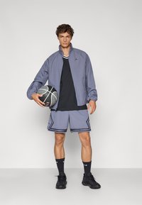 Chaqueta ligera morada, camiseta negra, pantalones cortos de malla grises con acentos negros, sosteniendo un balón de baloncesto, vistiendo zapatillas y calcetines negros.