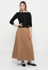 Woman standing with hand on hip, wearing black buttoned cardigan, white collar shirt, long brown skirt, gray socks, and dark red flats.