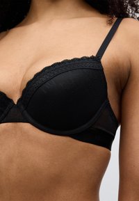 Μαύρο σουτιέν με δαντελένιο τελείωμα και δομημένο κύπελλο, διαθέτοντας υφασμάτινη υφή και ρυθμιζόμενους ιμάντες, με απαλό φινίρισμα.