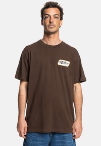 Brown short-sleeve t-shirt made from cotton, featuring a white and yellow logo patch on the left chest, loose fit, and crew neckline.
