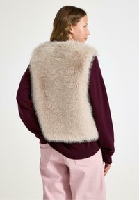 Beige nepbont vest over een diep bordeauxkleurige trui, met een geribbelde halslijn en een zachte textuur. Lichtroze broek maakt de outfit compleet.