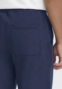 Temno modre trenirke iz mehkega blaga, s poudarkom na žepu zadaj in elastičnim pasom za udobje.