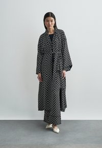 Černobílý dlouhý plášť s širokými rukávy, páskem v pase a ladící sukní. Hladká látka s geometrickým vzorem.