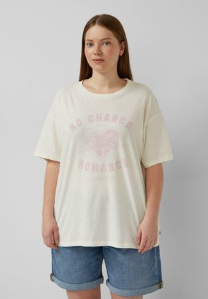 Młoda kobieta o długich brązowych włosach ubrana w biały, luźny T-shirt z wyblakłym sercem i napisem "No Chance Romance" oraz niebieskie szorty dżinsowe.