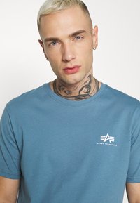 Blaues Baumwoll-T-Shirt mit Rundhalsausschnitt und kurzen Ärmeln, mit einem weißen Logo auf der linken Brustseite. Glatte Textur.