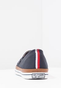 Navy blue casual slip-on shoe with brown sole, white and red vertical stripe on heel, and "Hilfiger Est. 1985" label on the back.
