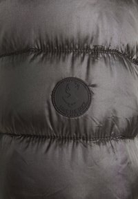 La giacca a vento nera presenta una texture liscia e lucida. Include un logo circolare nero "Save The Duck" su una sezione trapuntata.