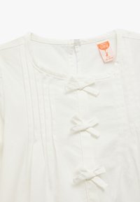 Prenda de algodón blanco con cuello redondo, textura de rayas verticales y un cierre frontal con tres pequeños lazos.