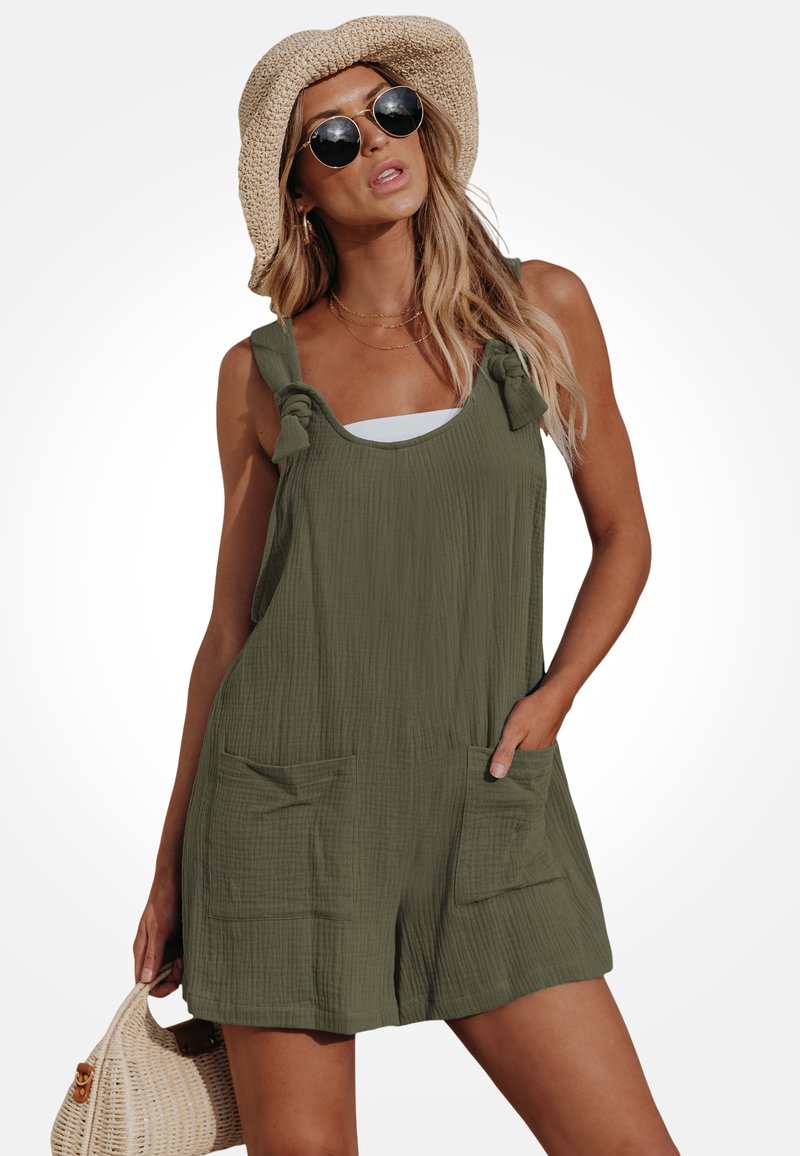 Vrouw die een olijfgroene mouwloze romper met voorzakken draagt, een ronde zonnebril, een strohoed en een geweven handtas vasthoudt.