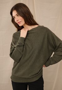 Naine, kellel on pikad pruunid juuksed, seljas oliivroheline kampsun ja mustad püksid, poseerib ühe käega kaela lähedal neutraalse tausta ees.