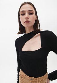 Sort ribbet langærmet top med en distinkt firkantet udsnit detalje på den øverste bryst. Glat tekstur og tætsiddende design.