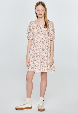 Młoda dziewczyna stojąca, ubrana w białą sukienkę w kwiaty z bufkami, kremowe skarpetki i beżowe trampki na tle jasnego, jednolitego tła.